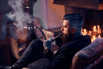 Attractive bearded man is making nice misty vapour while relaxing near fireplace and smoking hookah.