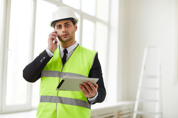Waist up portrait of Middle-Eastern engineer wearing hardhat speaking by phone while standing on construction site, copy space