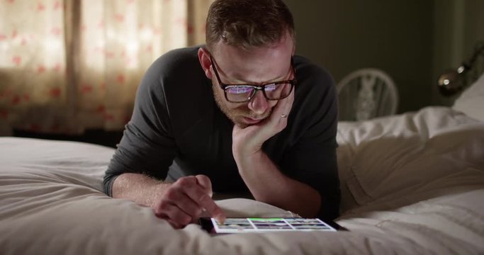 4K Tired man with internet addiction lying on his bed looking at digital tablet. Slow motion.