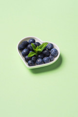 Fresh blueberries in heart shaped plate on pastel green background. Healthy breakfast concept