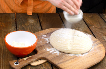 Making bread: sifting white flour on a formed loaf that is sitting on a pizza peel board.