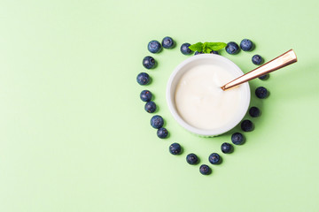 Bowl of natural yogurt and fresh blueberries in shape of heart on pastel green background. Healthy breakfast concept