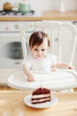 Fototapeta na wymiar Cute baby girl sitting in highchair, looking at piece of birthday cake with candle and trying to reach it with her tiny hand
