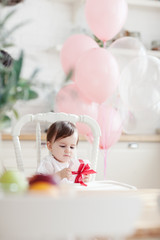 Adorable baby girl sitting in highchair in domestic kitchen and looking at birthday gift box in her hands. Room decorated with pink balloons