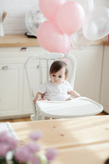 Fototapeta na wymiar Cute baby girl sitting in highchair decorated with balloons on her birthday and looking away thoughtfully