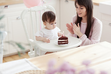 Happy young mother clapping hands supporting baby daughter sitting in highchair and trying to blow out her first candle on birthday cake