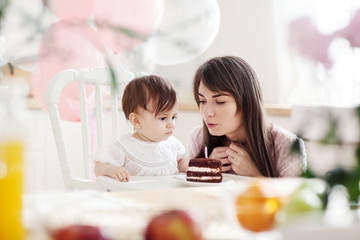 Obraz na płótnie Canvas Beautiful young mother teaching her baby daughter how to blow out candle on her first birthday cake