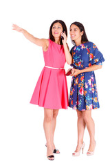 Two beautiful women, girlfriends, sisters in summer colored dress, pointing by finger and speak by mobile cell smartphone on isolated white background