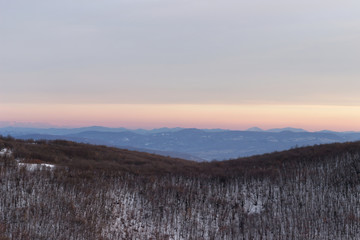 Beautiful scenic view of a forest on a snow covered mountain with distant mountain layers and soft colors of a sunset sky