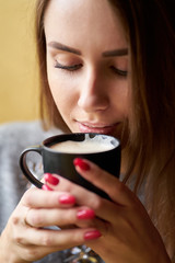 Attractive girl with long hair holding cup of cappuccino and drinking coffee in the cafe. Closeup portrait.