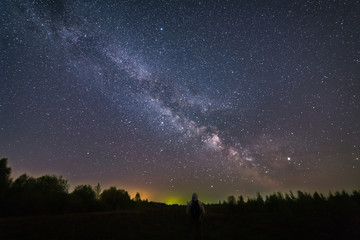 Naklejka na ściany i meble A man with a backpack standing at night, with his back to the camera, against the background of the Milky Way