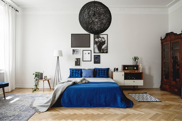 Real photo of navy blue bedroom in modern condo. Dark wooden china closet in the corner, small white dresser with vintage radio on it next to king size bed with velvet pillows and blanket.