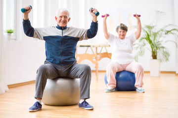 Two elderly people holding weights and sitting on exercising balls