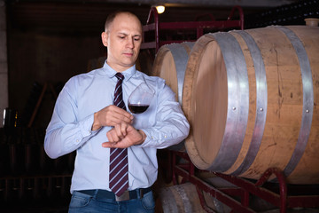 Confident male winemaker degusting red wine in wine cellar near bottles racks
