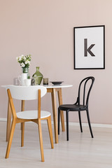 White jug, coffee cup and flowers in glass vase on white wooden dining table in spacious living room interior with poster on pastel pink wall