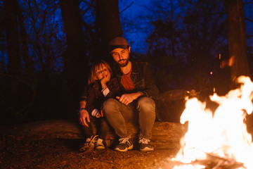 Happy father and his little son sitting together on the logs in front of a fire in a hike in the forest at the night.