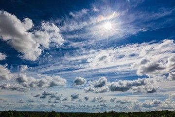 View of windy spring day with white clouds on blue sky and shining sun. Green forest on horizon.