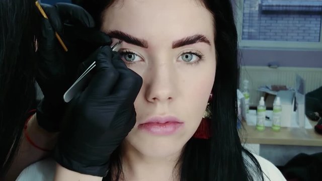 Cosmetologist's hand pulls out the beautiful patient's eyebrow with a pair of tweezers. Part of the eyebrow correction procedure. Professional face care in a beauty salon