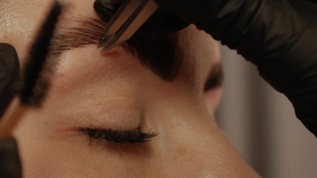 Close-up of a cosmetologist's hand pulls out the beautiful patient's eyebrow with a pair of tweezers. Part of the eyebrow correction procedure. Professional face care in a beauty salon