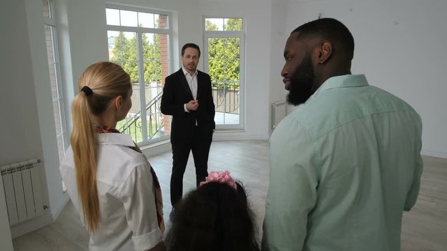 Back view of mixed race married couple standing in light spacious room with floor-to-ceiling window communicating with realtor and each other. Real estate agent showing house to new homeowners