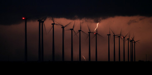 The silhouette of spinning wind turbines is backlit by a bolt of lightning in a thunderstorm at night.