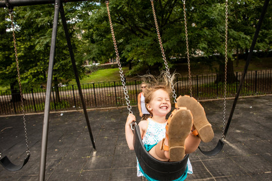 A toddler aged girl swinging and having fun outdoors at a park with her mother.