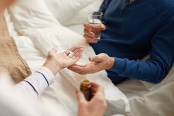 Closeup of unrecognizable senior man lying on bed and taking pills with caring wife or nurse , copy space