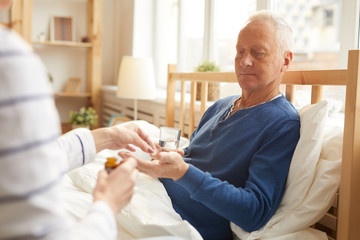 Portrait of senior man lying in bed and taking pills with caring wife or nurse , copy space