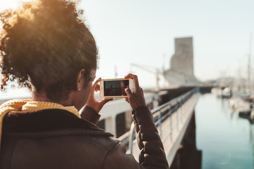 View from behind of a curly-hair female in leather demi-season coat photographing in a landscape mode a cityscape with quayside, buildings, yachts, and boats using the camera of her smartphone