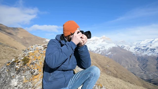 Portrait of a bearded traveler photographer in sunglasses and a cap sits on a rock with mirror camera in his hands and take a picture against the backdrop of mountains