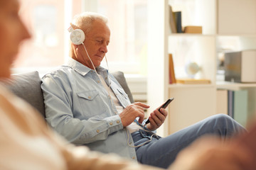 Portrait of modern senior man wearing headphones using smartphone sitting on sofa at home, copy space