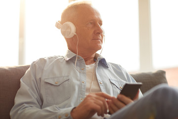 Portrait of modern senior man wearing headphones using smartphone sitting on sofa at home lit by...