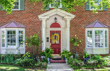 Entrance with columns and bay windows of beautiful two-story brick house with pots of flowers and yellow climbing roses and headplanters and ferns frame by Maple tree limbs