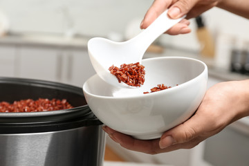 Woman putting brown rice into bowl from multi cooker in kitchen, closeup