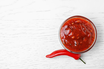 Bowl of hot chili sauce with red pepper on white wooden background, top view. Space for text