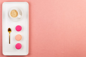 Pink french macarons or macaroons, coffee cup and golden spoon on a rectangle white plate over a pink tablecloth background with copy space.