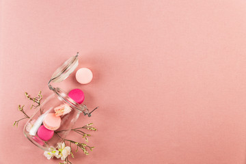 Pink french macarons or macaroons, falling out of a glass jar with white flowers over a pink tablecloth background with copy space.