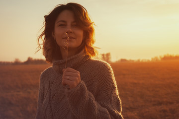 Portrait joyful young woman   brunette in brown knit sweater made of natural wool and jeans holding a spike and smiles mysteriously and enjoy day on field. stylish hipster woman.
