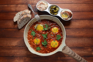 Shakshuka , middle eastern traditional homemade breakfast- fried eggs, onion, bell pepper, tomatoes and parsley in a pan on a wooden background served with hummus