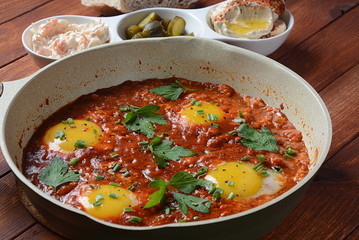 Shakshuka , middle eastern traditional homemade breakfast- fried eggs, onion, bell pepper, tomatoes and parsley in a pan on a wooden background served with hummus