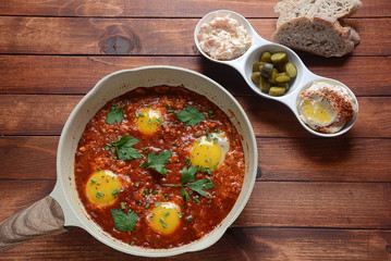 Shakshuka , middle eastern traditional homemade breakfast- fried eggs, onion, bell pepper, tomatoes and parsley in a pan on a wooden background served with hummus