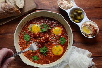 Shakshuka , middle eastern traditional homemade breakfast- fried eggs, onion, bell pepper, tomatoes and parsley in a pan on a wooden background served with hummus