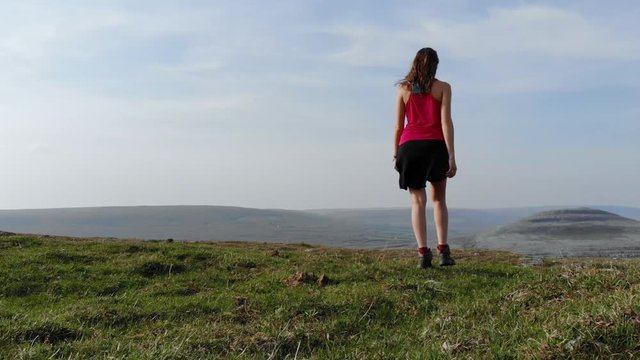 A Women Hiker Walks Away From The Camera On Top Of A Hill With Views Of Grass Fields  And Rocky Hills In The Burren National Park In Ireland.
