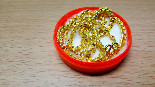 The Gold Necklace Is Placed In A Red Plastic Box On The Wooden Table.