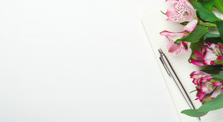 Notepad with steel pen on a white wooden background with flowers. Minimal composition, home women's office, copy space, top view, flat lay.