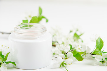 Face cream in white jar on a white background with white small flowers of an apple tree. Concept natural cosmetics, organic beauty, delicate floral composition.