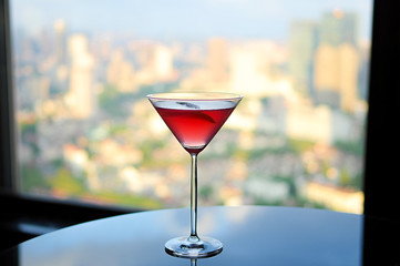 Red cocktail with fruit in martini glass on clean lounge table in daylight with cityscape as background