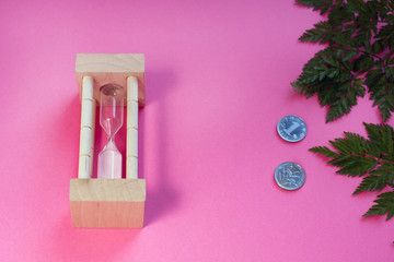 Hourglass with coins and fern leaves on pink background
