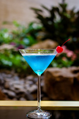 Blue lagoon cocktail in martini glass with red cherry at indoor garden with plants