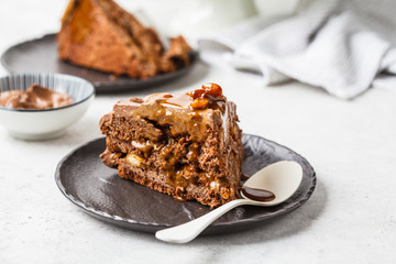 Piece of homemade snickers cake with chocolate cream and caramel on a black saucer, white...